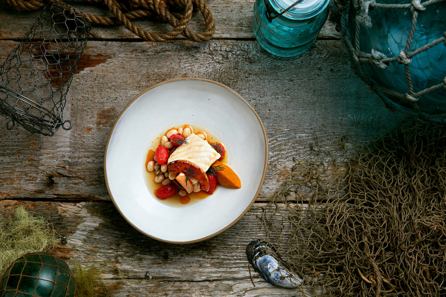 A rustic wooden table displays a plate of fish fillet garnished with white beans, tomatoes, and a sauce. Around the plate are fishing nets, a shell, glass buoy, and coiled rope, evoking a coastal theme.