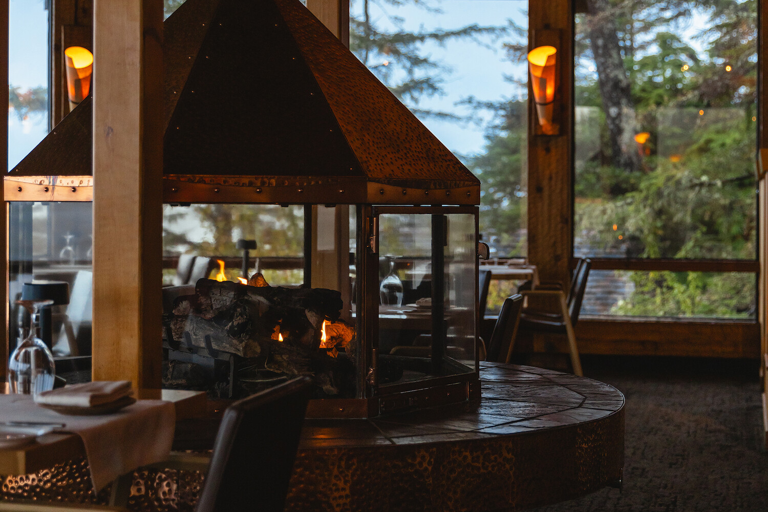 Cozy restaurant interior featuring a central, glass-enclosed fireplace with a warm glow. Large windows reveal a coastal outdoor view, and tables are set with white cloths, offering a charming dining atmosphere.